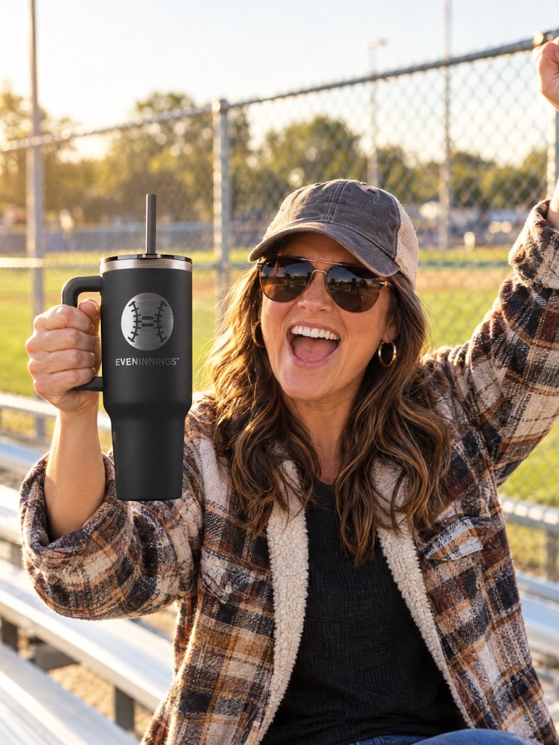 A mom cheering in the bleachers, holding up the 40 oz Tumbler in black with the Even Innings ball mark and name laser-etched on the side.